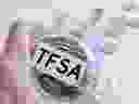 Hand holding glass jar with many coins and TFSA word over a marble counter top.  