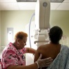 An African American woman is shown getting a mammogram. She is photographed from behind, so we see the back of her head and body as she stands facing a large X-ray machine. A health care professional wearing pink scrubs positions the woman in the machine.