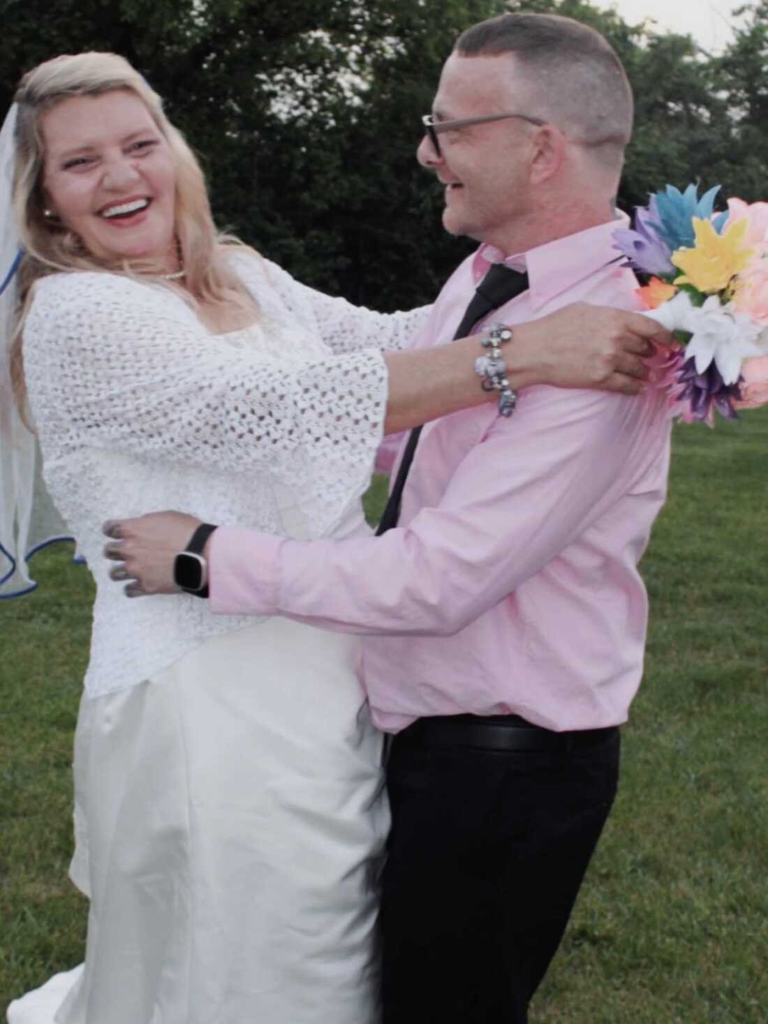 TJ Hoover danced with his sister, Donna, on her wedding day in May 2023. Donna has long blond hair and is wearing a white wedding dress. TJ is wearing a pink dress shirt and black pants. She has a bouquet in her hands. They are outside, dancing on green grass near trees.