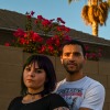 Samantha and Ariane Buck are a young couple standing in front of a house with a high-walled garden. Bright red flowers peek over the wall above their heads.