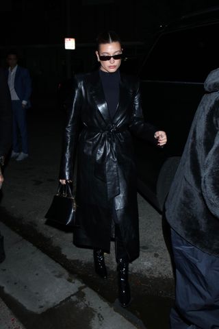 Hailey Bieber wearing a black Khaite leather trench coat with a turtleneck, sunglasses, and black Saint Laurent clasp bag.